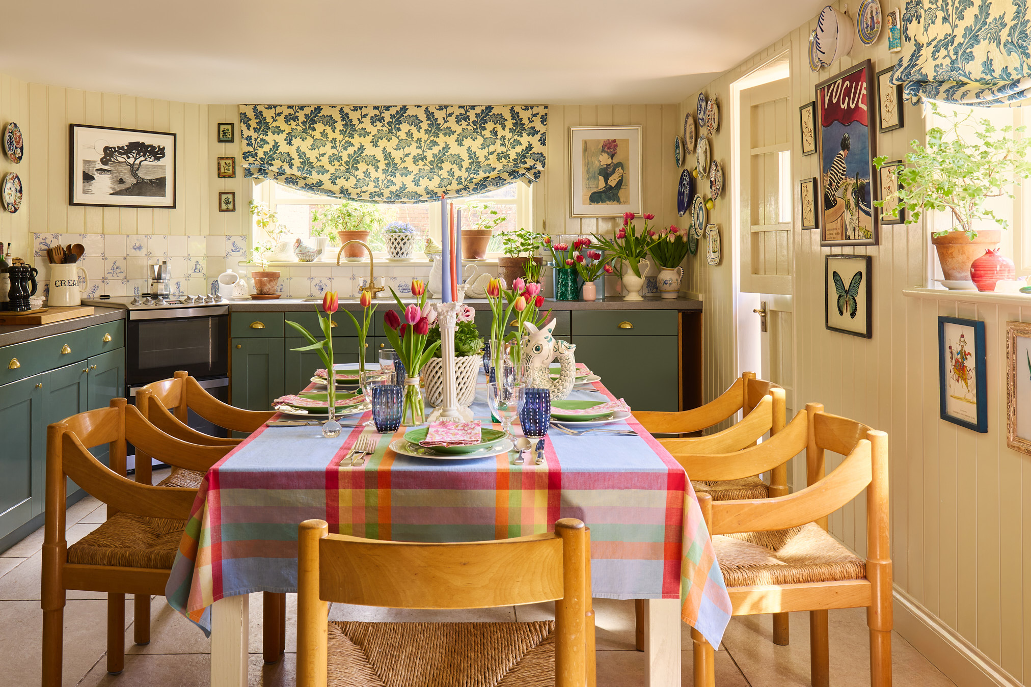 Kitchen with green painted cabinets, buttermilk painted panelling, floral gathered blinds, wooden table set with colourful tablecloth and tulips in a Hampshire cottage