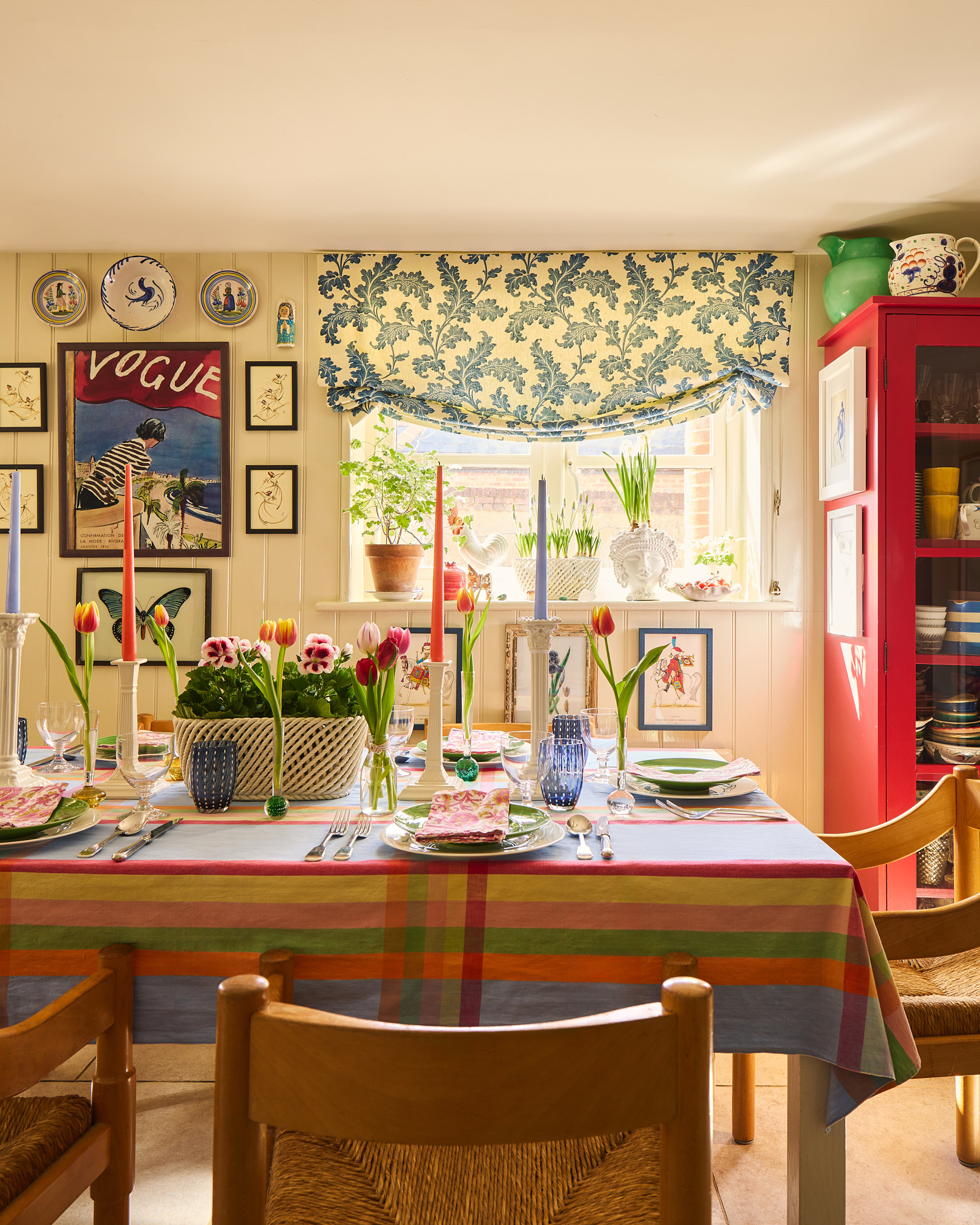 Kitchen with red painted dresser, buttermilk painted panelling, floral gathered blinds, wooden table set with colourful tablecloth and tulips in a Hampshire cottage